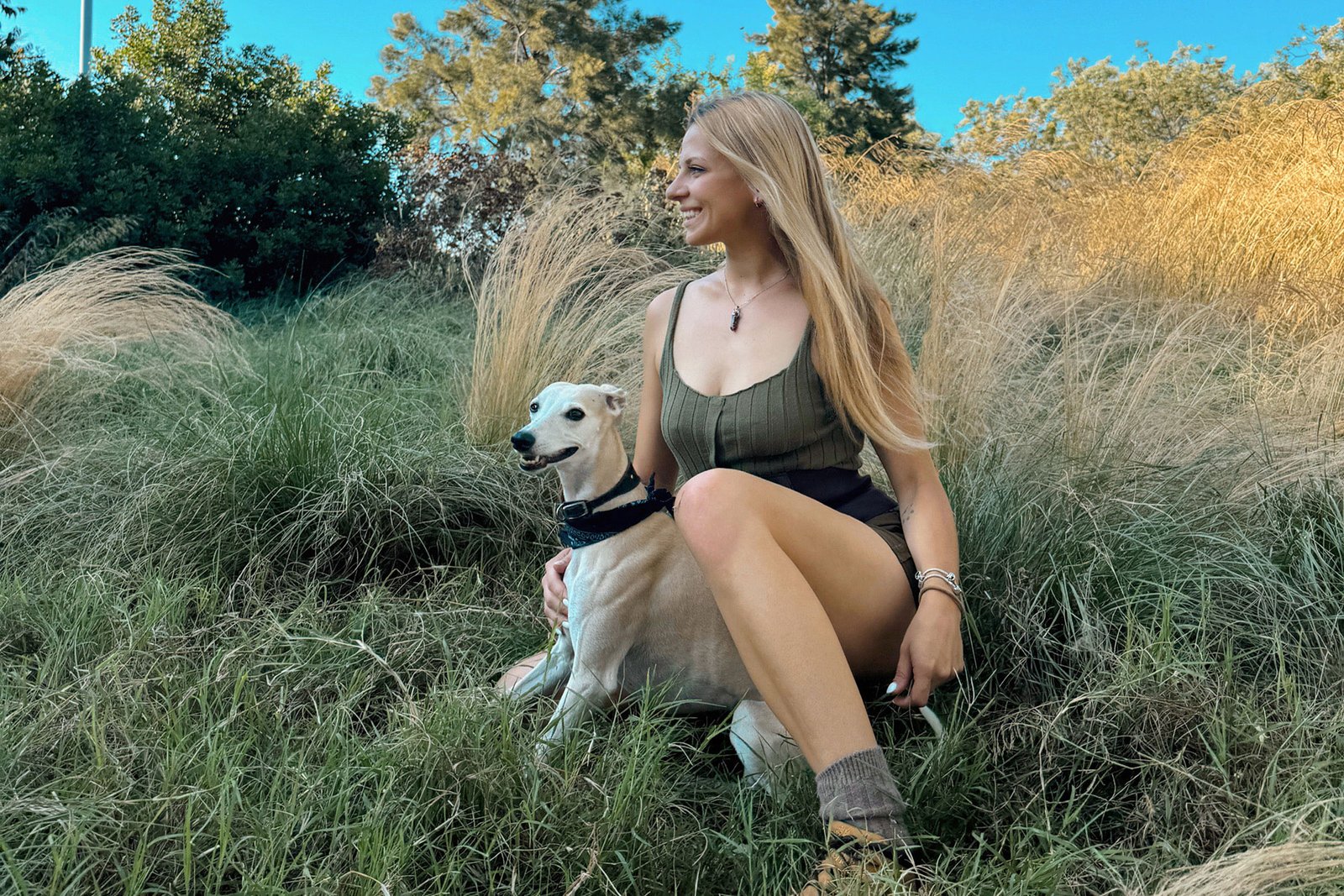 Joven sonriendo junto a su perro galgo en un campo al atardecer.